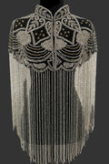 A black embellished shrug or cape with intricate silver beadwork, sequins, and long hanging fringe, displayed on a black dress form against an indoor wall.
