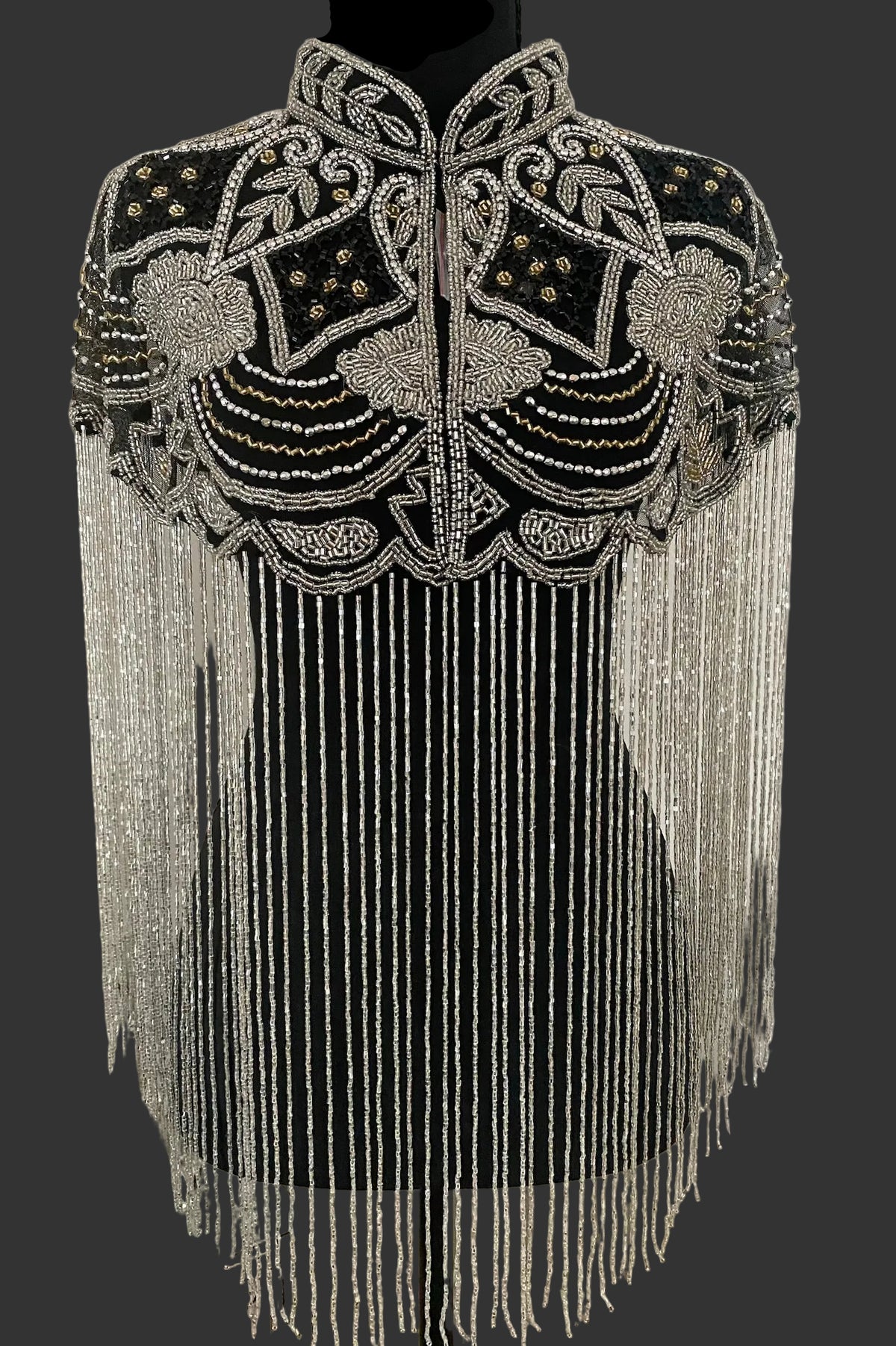 A black embellished shrug or cape with intricate silver beadwork, sequins, and long hanging fringe, displayed on a black dress form against an indoor wall.