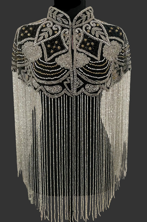 A black embellished shrug or cape with intricate silver beadwork, sequins, and long hanging fringe, displayed on a black dress form against an indoor wall.